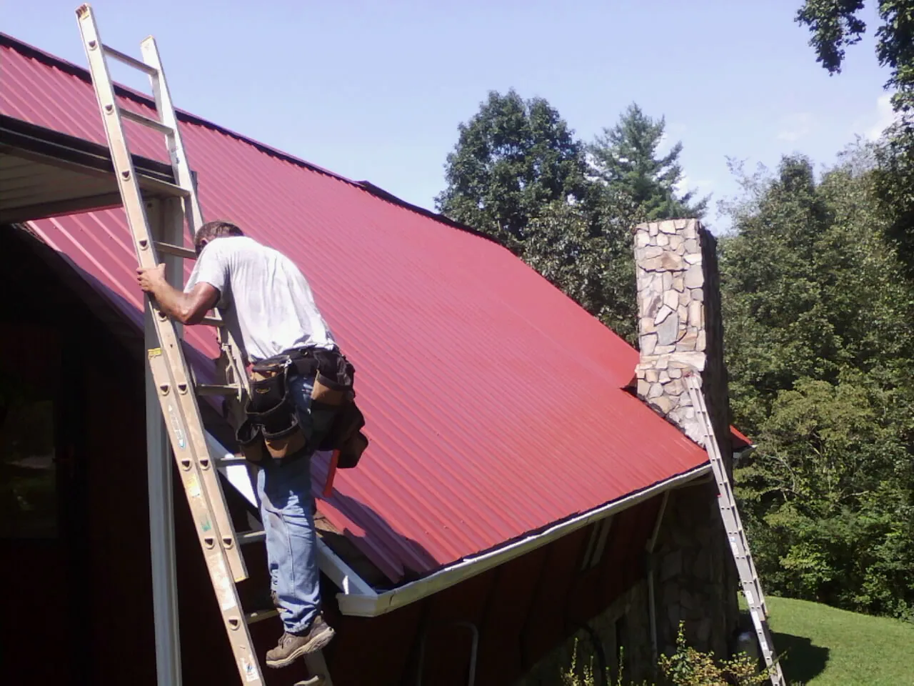 Roofer on a ladder beside a red metal roof and stone chimney — Soffit & Fascia Repair by Roofing Brothers in Luna Pier